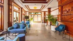 Un vestíbulo amplio con techo alto, ventanas grandes y vistas a palmeras. Hay varias zonas de estar con sofás y sillones azules.