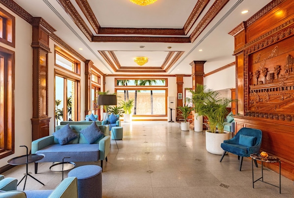 A spacious lobby with a high ceiling, large windows, and a view of palm trees. There are several seating areas with blue sofas and armchairs.