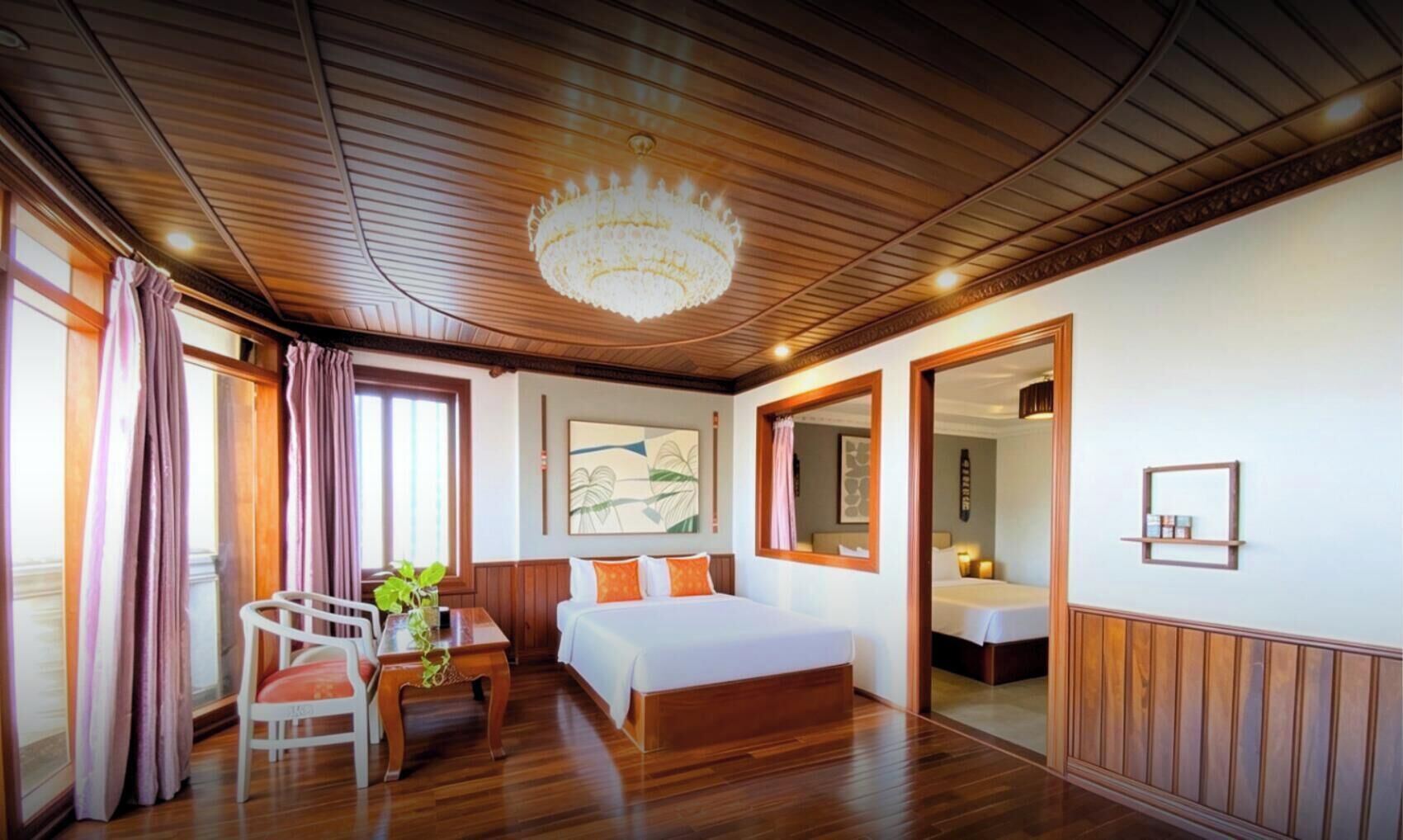 A hotel room with a wooden ceiling, a chandelier, a bed, a desk with a chair, and a window with curtains.