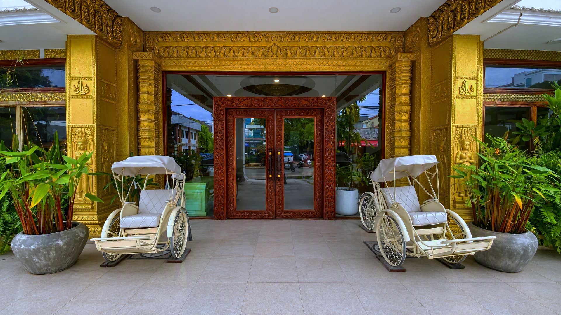 A building entrance with ornate golden pillars, a glass door, and two rickshaws on either side.