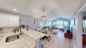 Eine moderne Küche mit Marmorarbeitsplatte und integriertem Spülbecken, ein Essbereich mit Glastisch sowie ein Wohnzimmer mit Sofa und Fernseher.