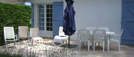 Eine Terrasse mit weißen Plastikstühlen und einem Tisch, einem blauen Sonnenschirm und einem Gebäude mit blauen Fensterläden.