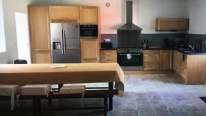 A kitchen with wooden cabinets, a built-in oven, and a dining table with a bench. - Gîte 12 Personnes (Brem-sur-Mer)