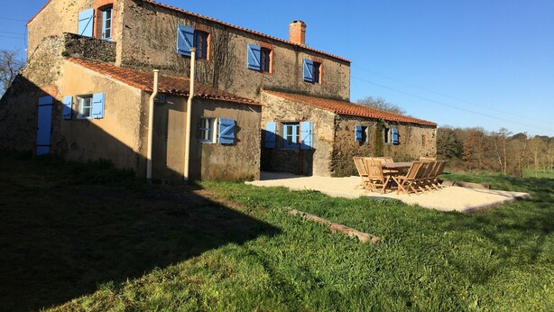 A rustic two-story house with a tiled roof and blue shutters, surrounded by a grassy area with a wooden table and chairs. - Gîte 12 Personnes (Brem-sur-Mer)