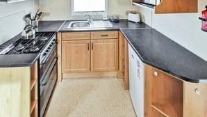 A compact kitchen with wooden cabinets, a black countertop, a stove, a sink, and a microwave. - Comfortable caravan with one double and two twin. (Fishguard, Pembrokeshire)