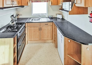 A compact kitchen with wooden cabinets, a black countertop, a stove, a sink, and a microwave. - Comfortable caravan with one double and two twin. (Fishguard, Pembrokeshire)