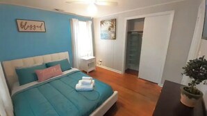 A bedroom with a bed, a bedside table, a nightstand, a window with curtains, a door, and a potted plant.