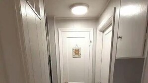 A narrow hallway with white walls and a wooden floor, leading to a door with a small picture frame.