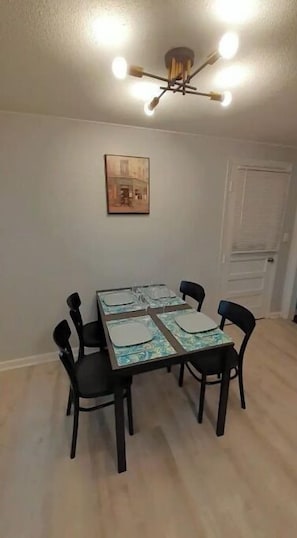 Uma sala de jantar com uma mesa posta para quatro pessoas, rodeada por cadeiras, um lustre no teto e um quadro na parede.