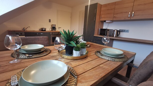 Une table à manger dressée avec des assiettes, des couverts et des verres à vin, avec une cuisine en arrière-plan.