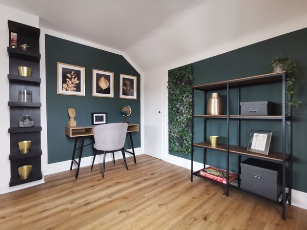 Une pièce avec un bureau en bois, une chaise, un globe terrestre, des tableaux encadrés, une bibliothèque et un mur vert orné de motifs végétaux.