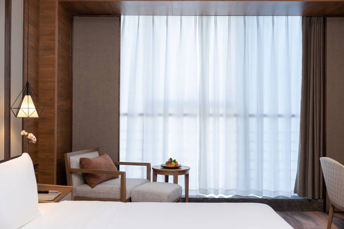 Ein Hotelzimmer mit Bett, Sessel, kleinem Tisch mit Früchten und großem Fenster mit durchsichtigen Vorhängen.
