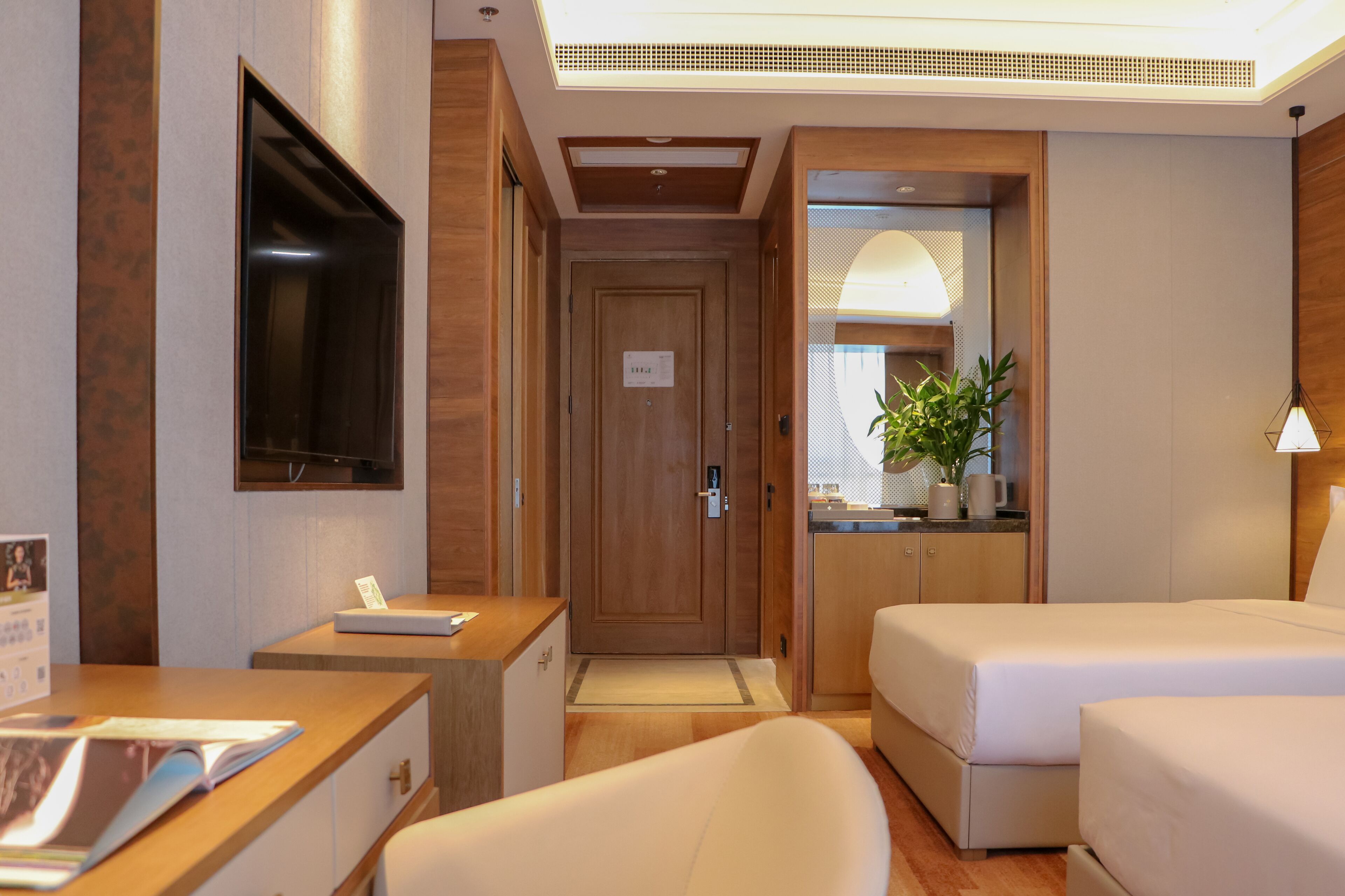 A hotel room with a desk, two beds, a TV, a plant, and a bathroom visible through an open door.