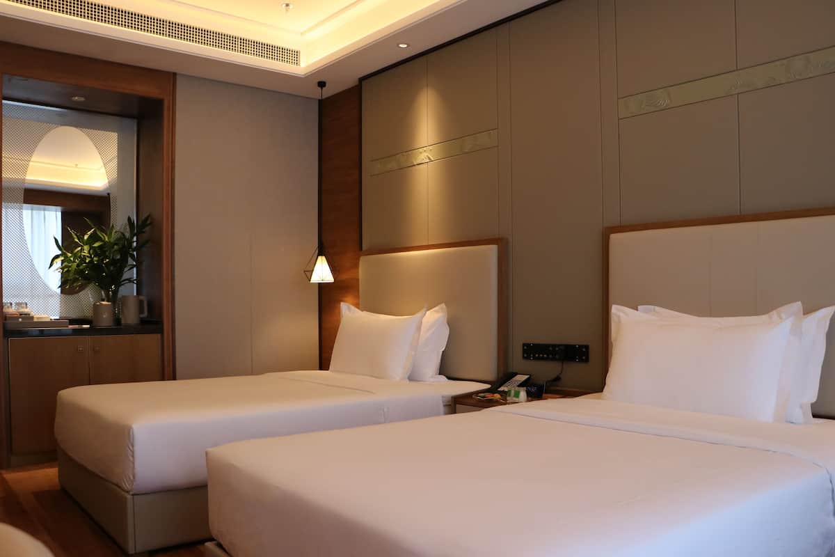 Ein Hotelzimmer mit zwei Einzelbetten, einem Nachttisch mit Lampe, einem kleinen Schreibtisch mit Telefon und einem Spiegel mit Pflanze.