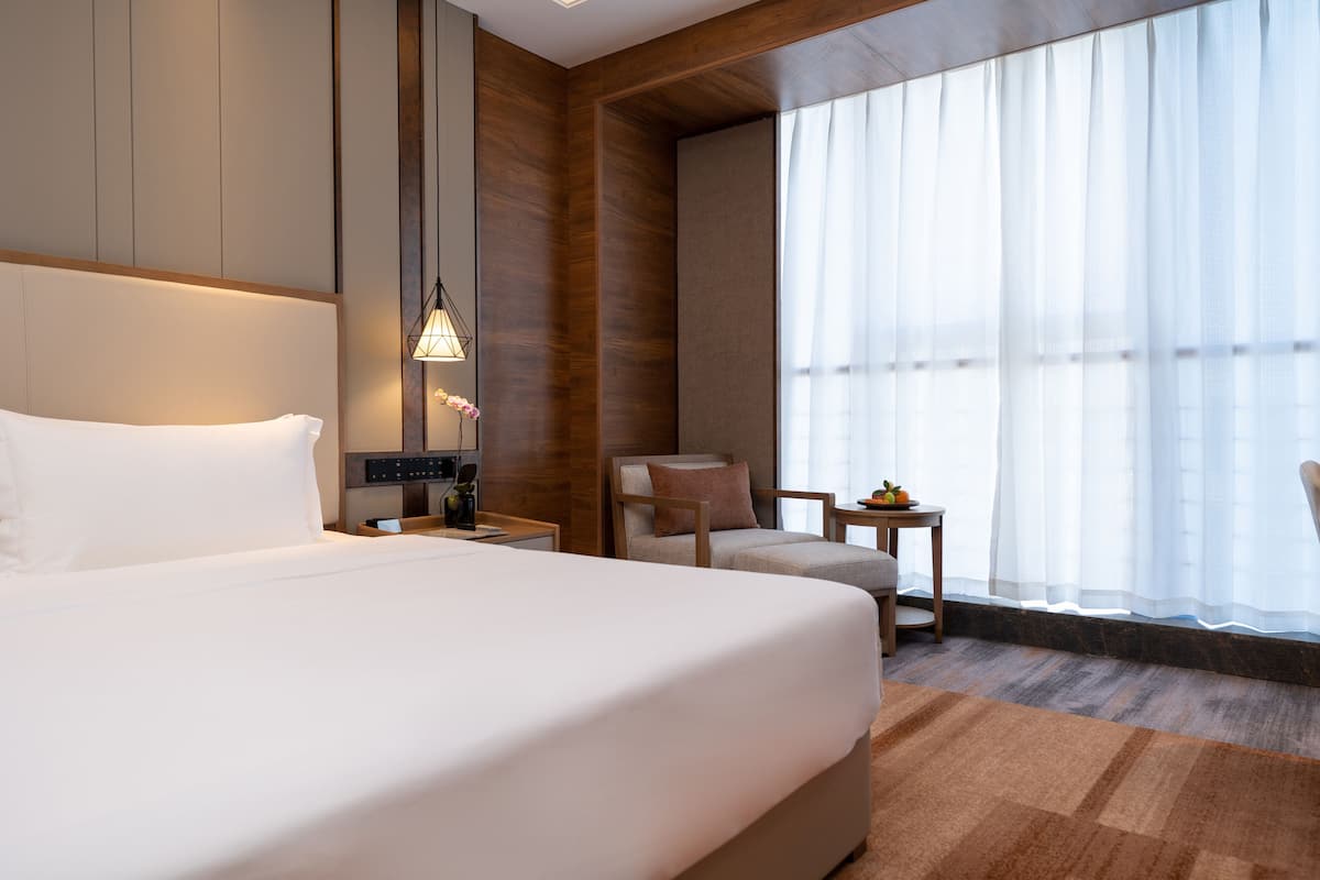 Ein modernes Hotelzimmer mit einem großen Bett, einem Schreibtisch mit Lampe, einem Sessel und Blick ins Freie durch transparente Vorhänge.