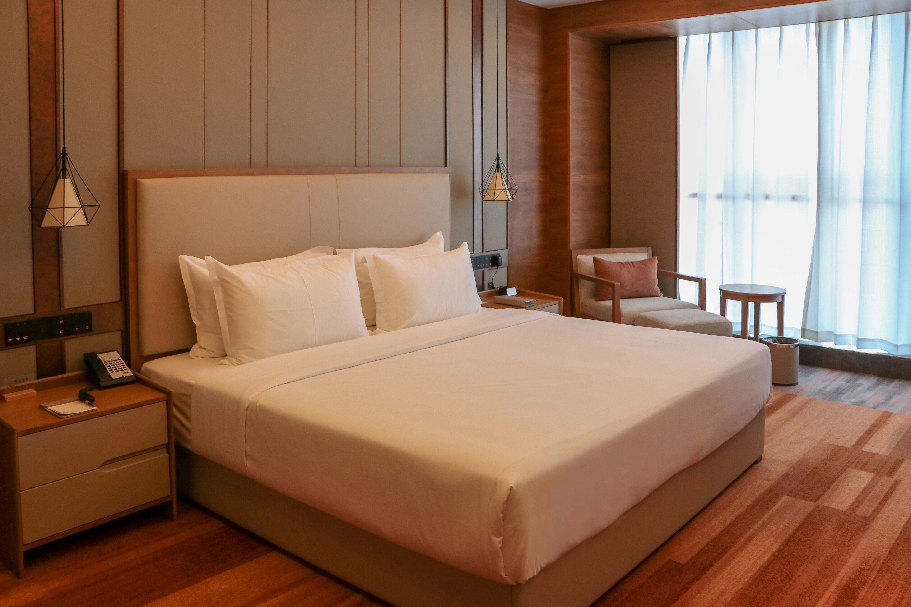 A hotel room with a large bed, bedside tables, a nightstand, a chair, a small table, and a window with curtains.