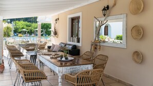 Un espacio de comedor al aire libre con techo, mobiliario de mimbre, una mesa central con un centro decorativo y vistas a una piscina y follaje en el fondo.