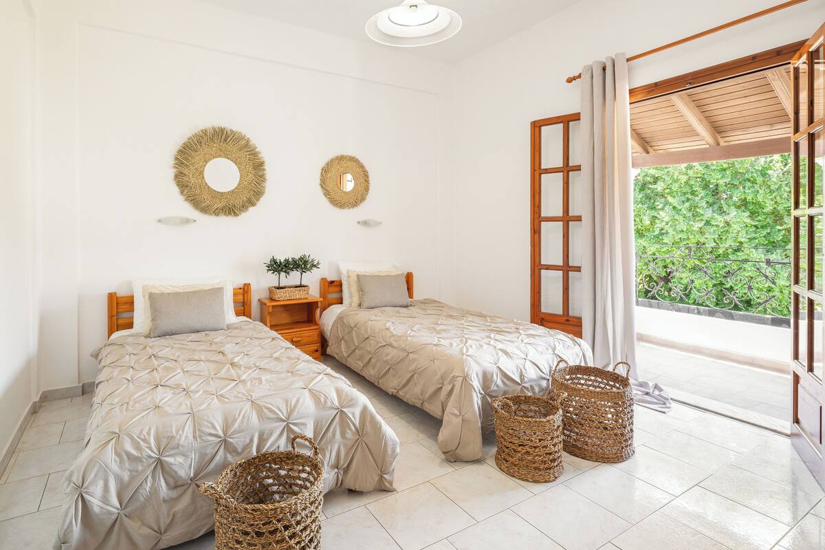 A bedroom with two beds, a wooden nightstand, wicker baskets, and a view of greenery through an open window.