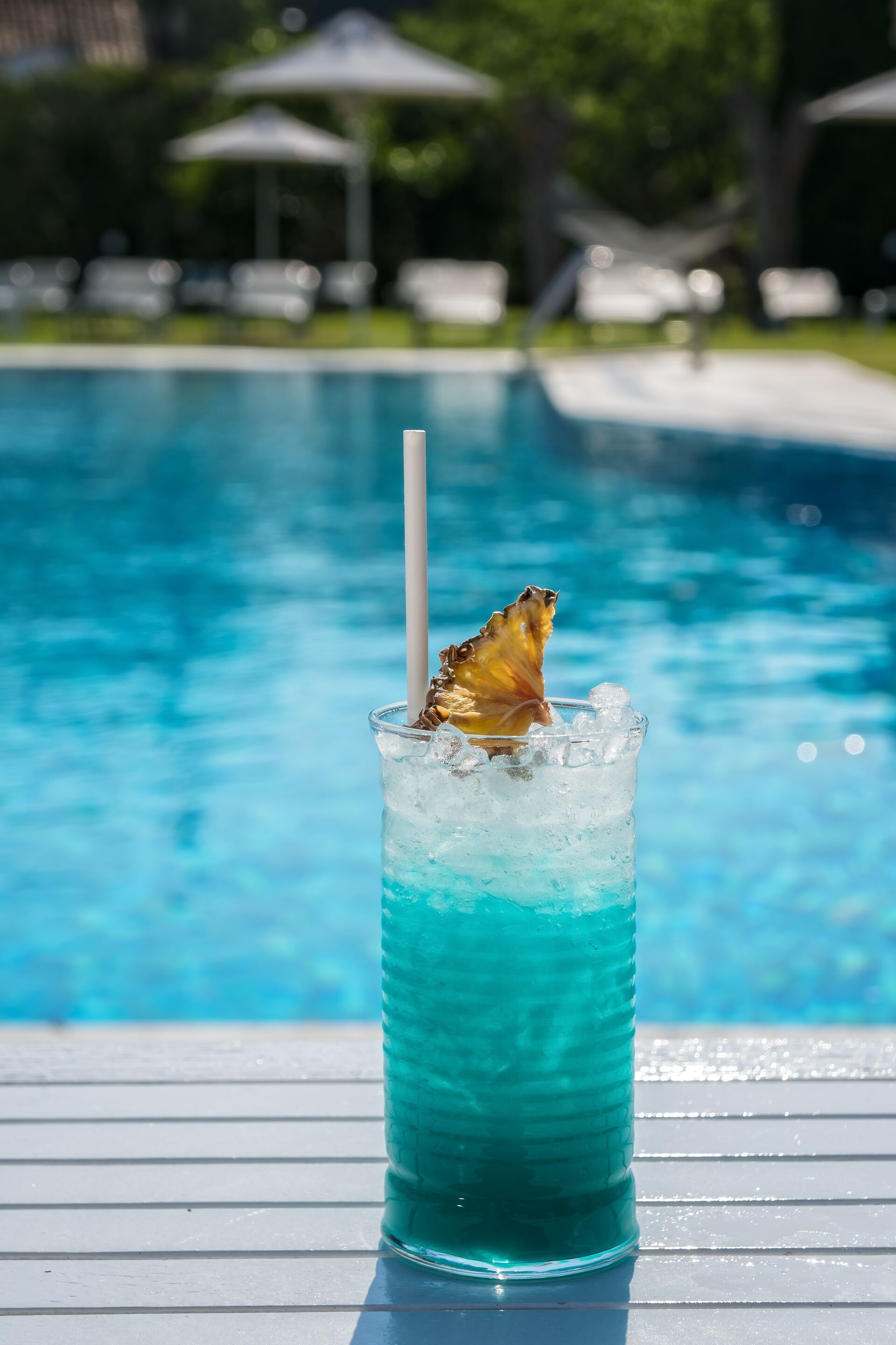 A glass of blue beverage with a straw and a pineapple slice on a poolside table.