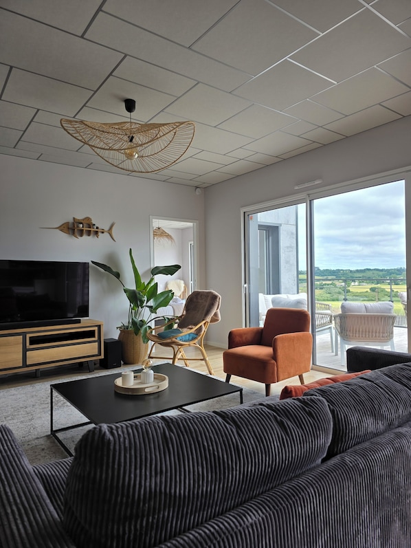 A modern living room with a large flat-screen TV, a sofa, a coffee table, and a balcony with a view of the countryside.