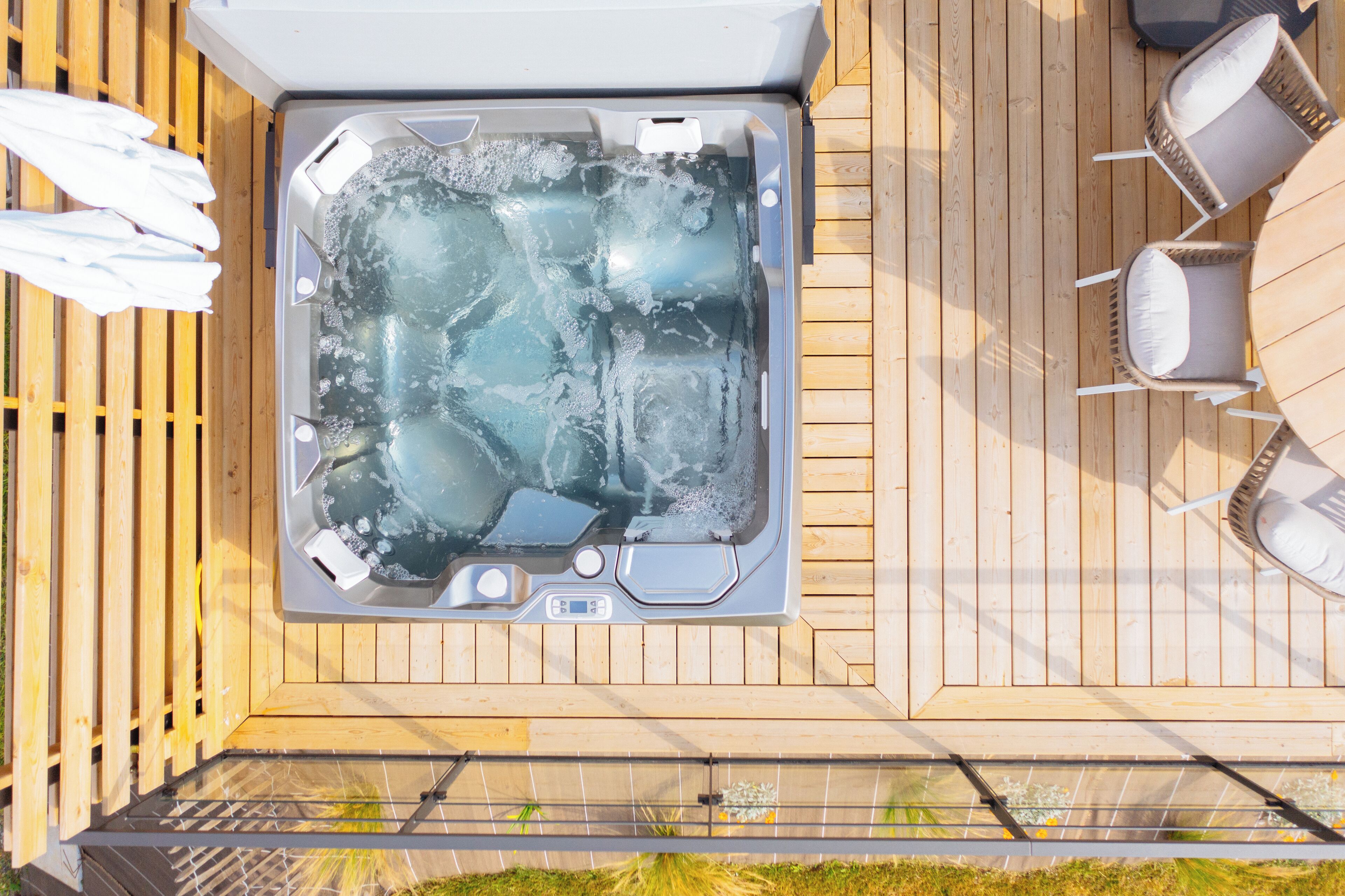 Un jacuzzi en una terraza de madera con mobiliario exterior blanco y barandilla de vidrio.