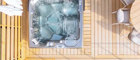 Un jacuzzi en una terraza de madera con mobiliario exterior blanco y barandilla de vidrio.