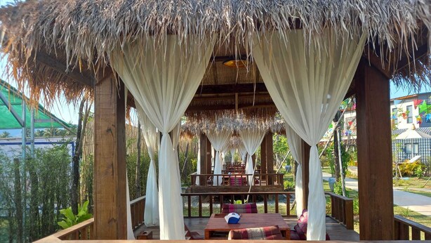 A wooden gazebo with a thatched roof, white curtains, and a wooden deck with seating. - Kampot Tropical Village (Kampot)