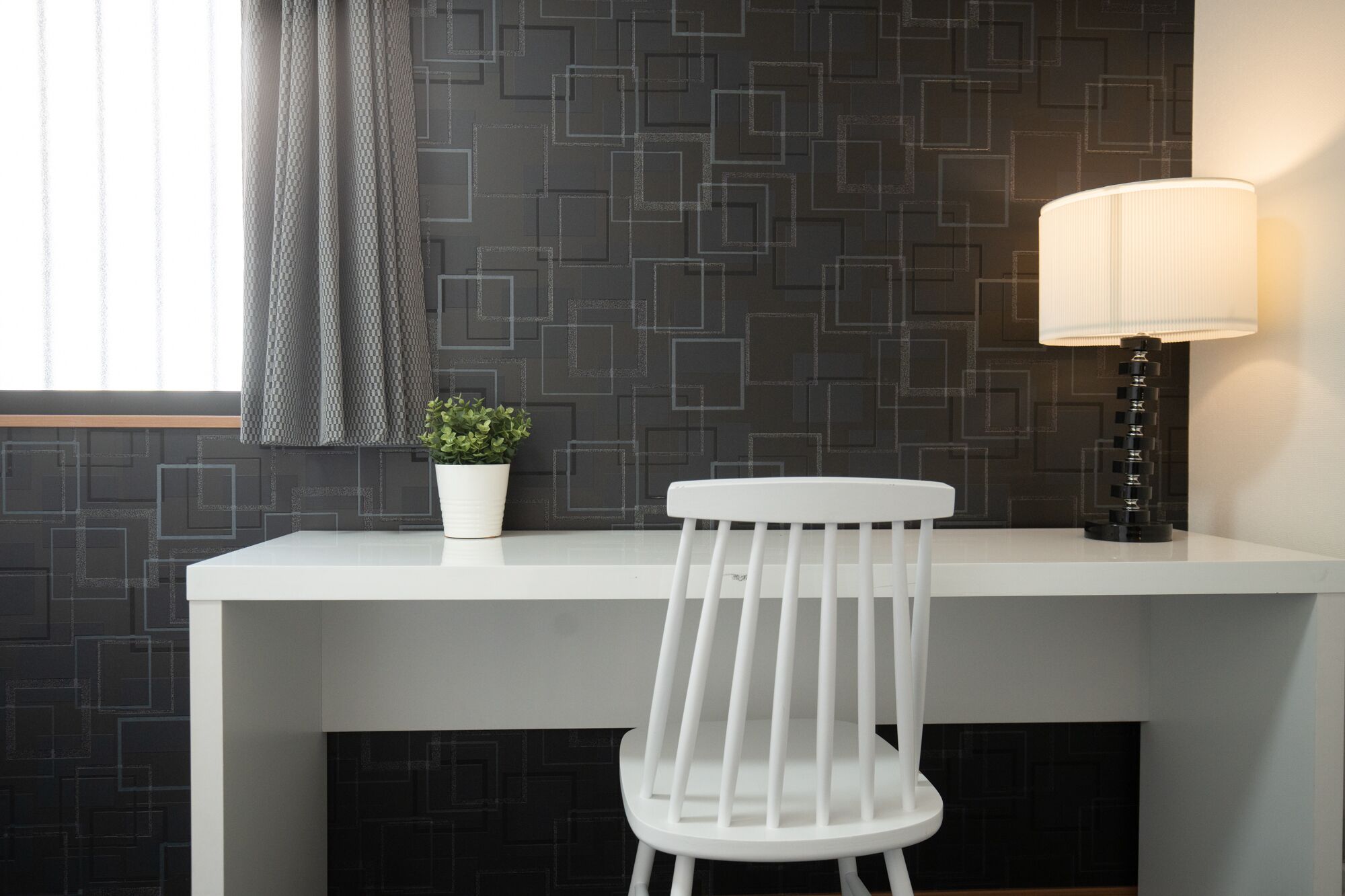 Ein moderner Schreibtisch mit weißem Stuhl, einem Topfpflanzen und einer Stehlampe vor einer Wand mit geometrischem Muster.