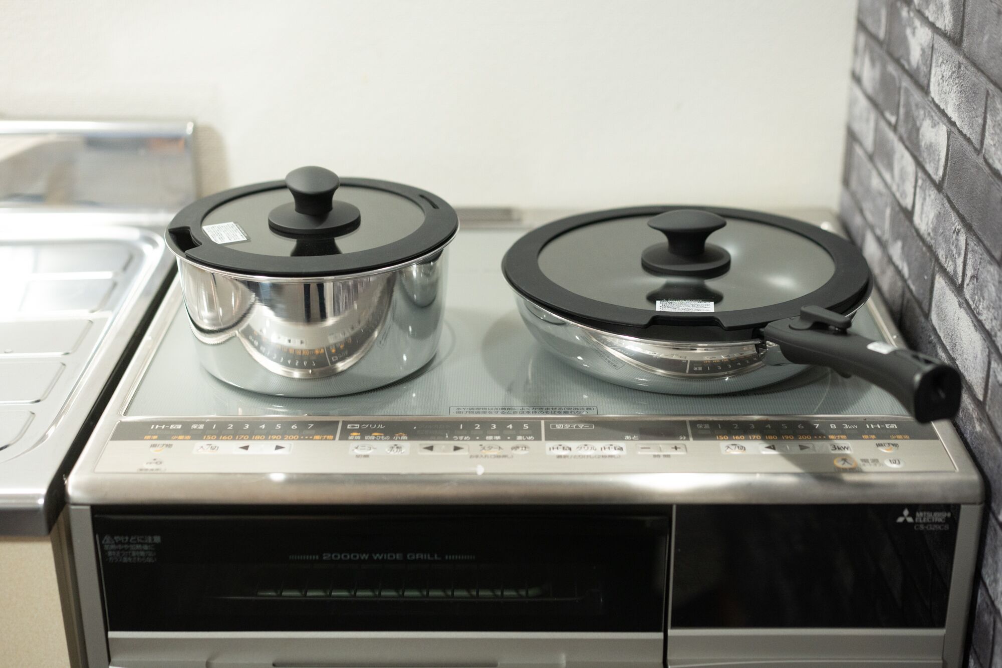 A 2000W wide grill stove with two pots, a control panel, and a brick wall in the background.
