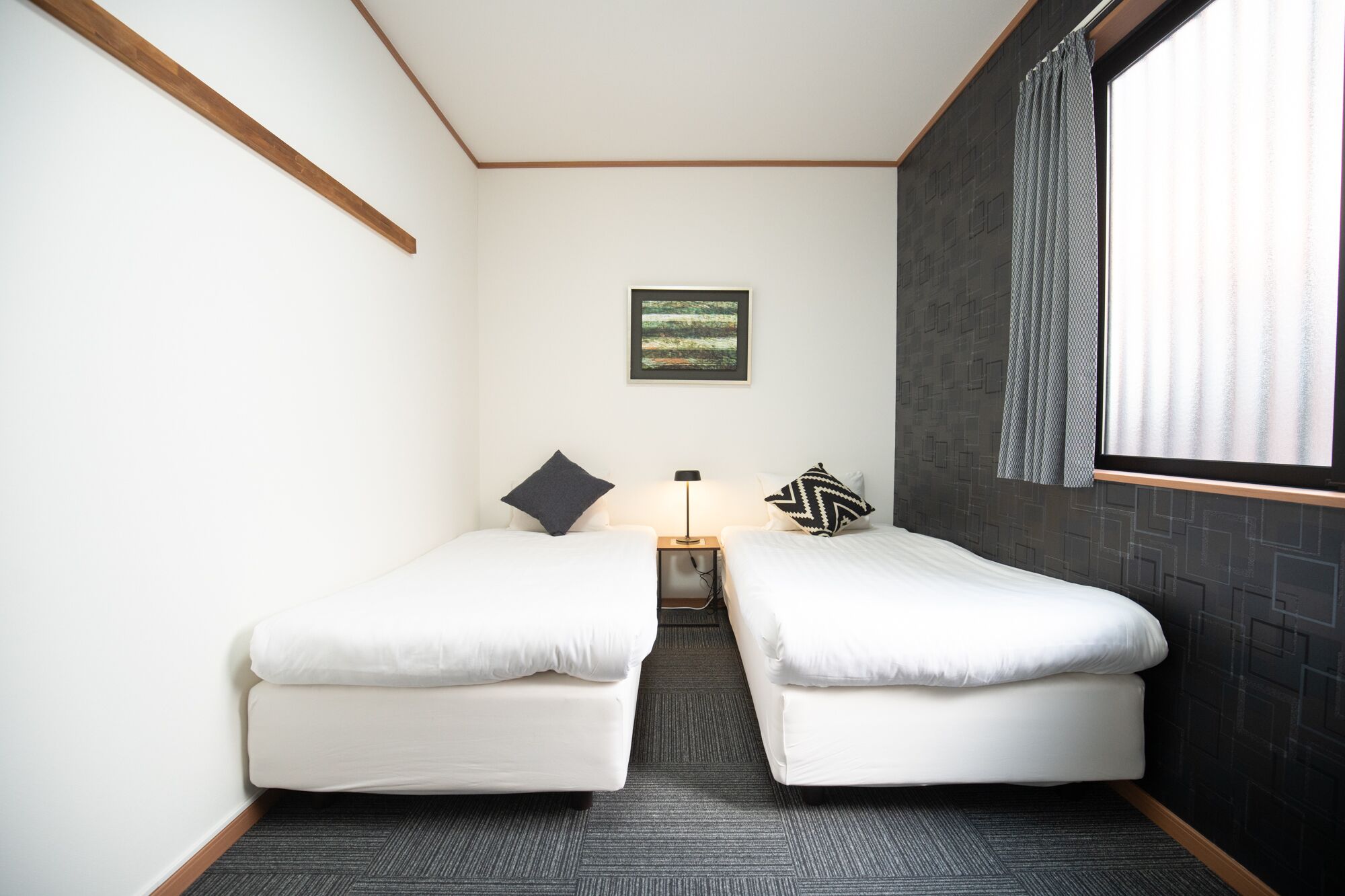 A small, modern room with two single beds, a nightstand with a lamp, a framed picture on the wall, and a window with curtains.