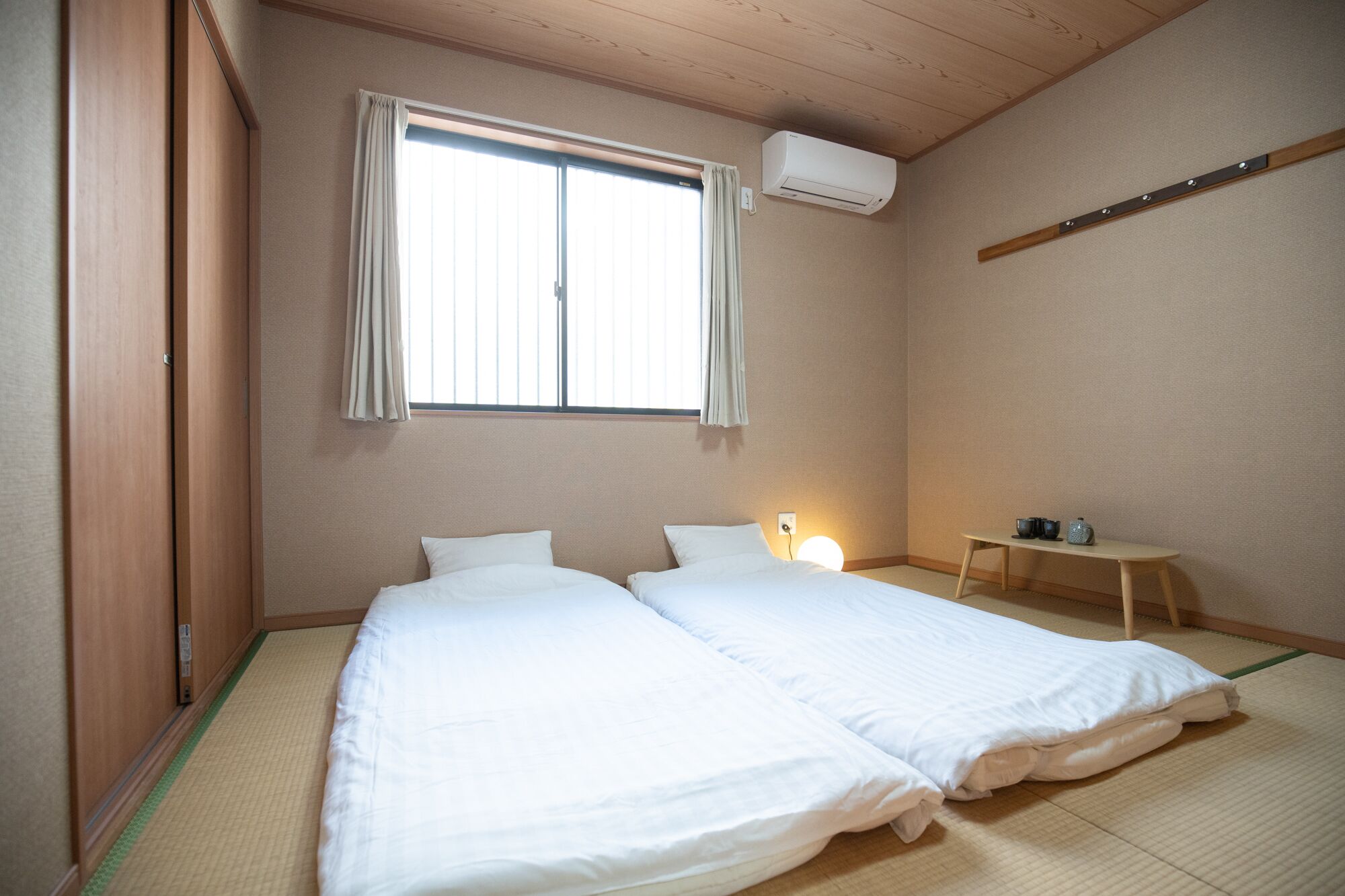 A room with two beds, a window with curtains, an air conditioner, a small table with cups, and tatami flooring.