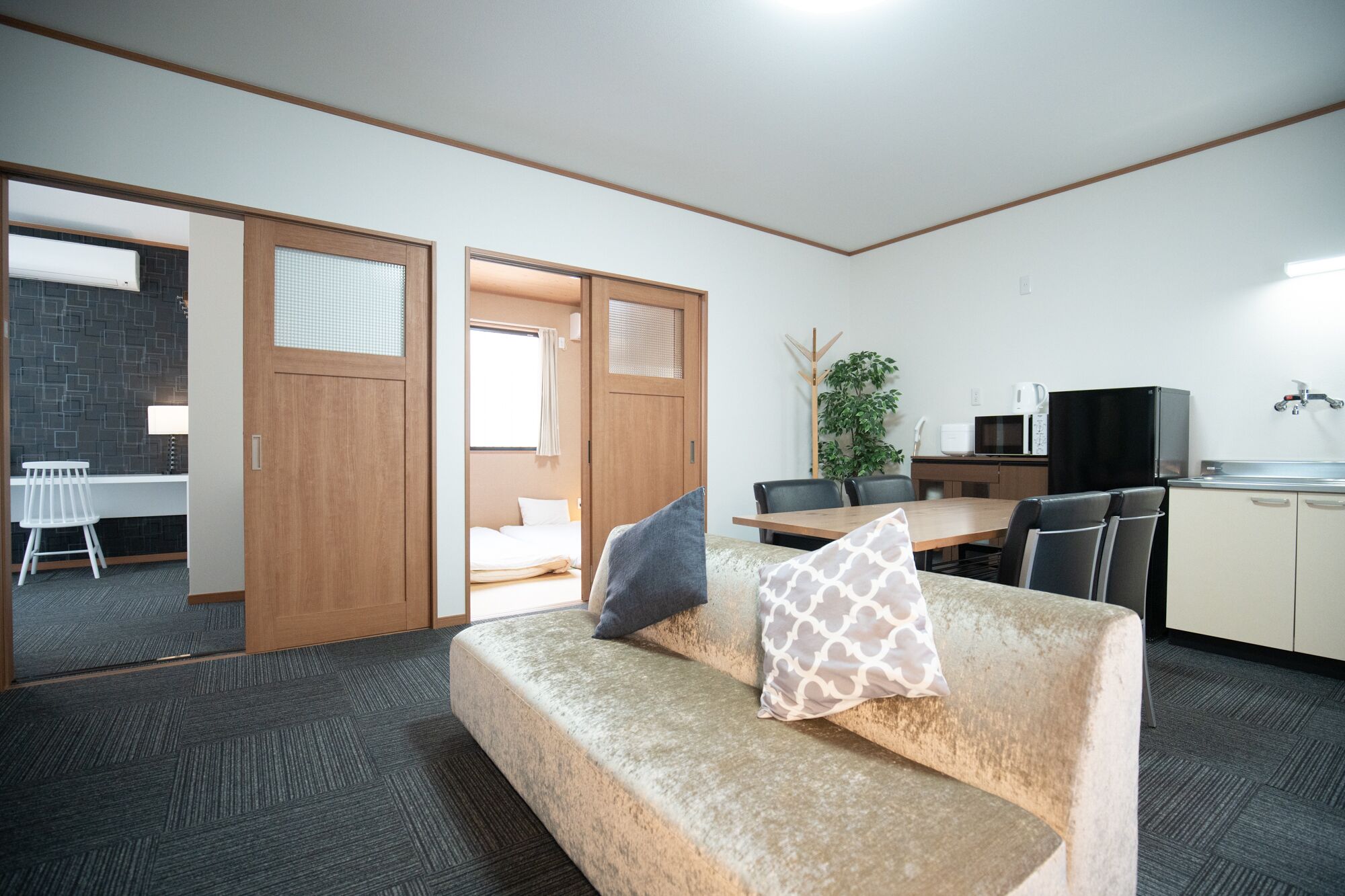 A modern hotel room with a sofa, a dining table, a kitchen area, and a sliding door leading to a balcony.