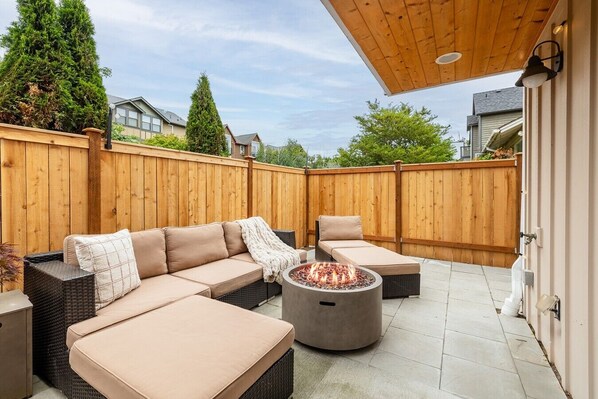 An outdoor patio with a wooden deck, a beige sofa set, a round table with a fire pit, and a pergola overhead.