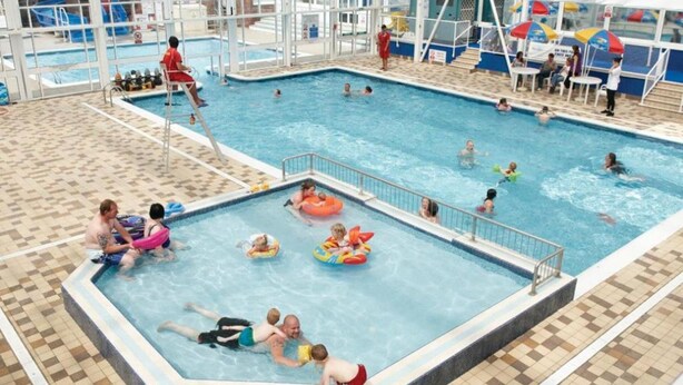 Une piscine intérieure dotée de plusieurs couloirs de nage, d’un poste de surveillance pour les sauveteurs et d’un espace aménagé au bord du bassin avec des parasols.