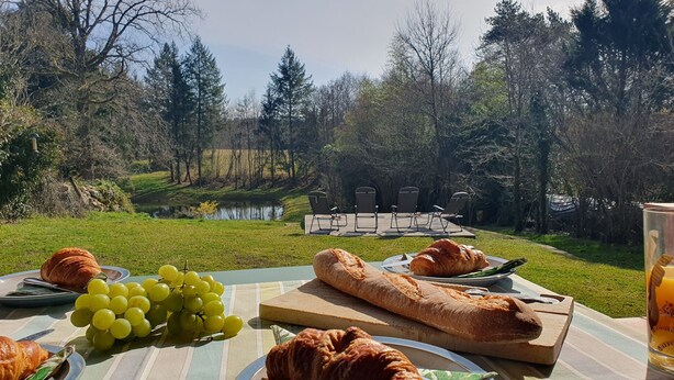 An outdoor dining setup with croissants, grapes, and a baguette on a table, overlooking a lake and forested area. - Maison de l'olivier - Rural Comfort & Relaxation (Champniers-et-Reilhac)