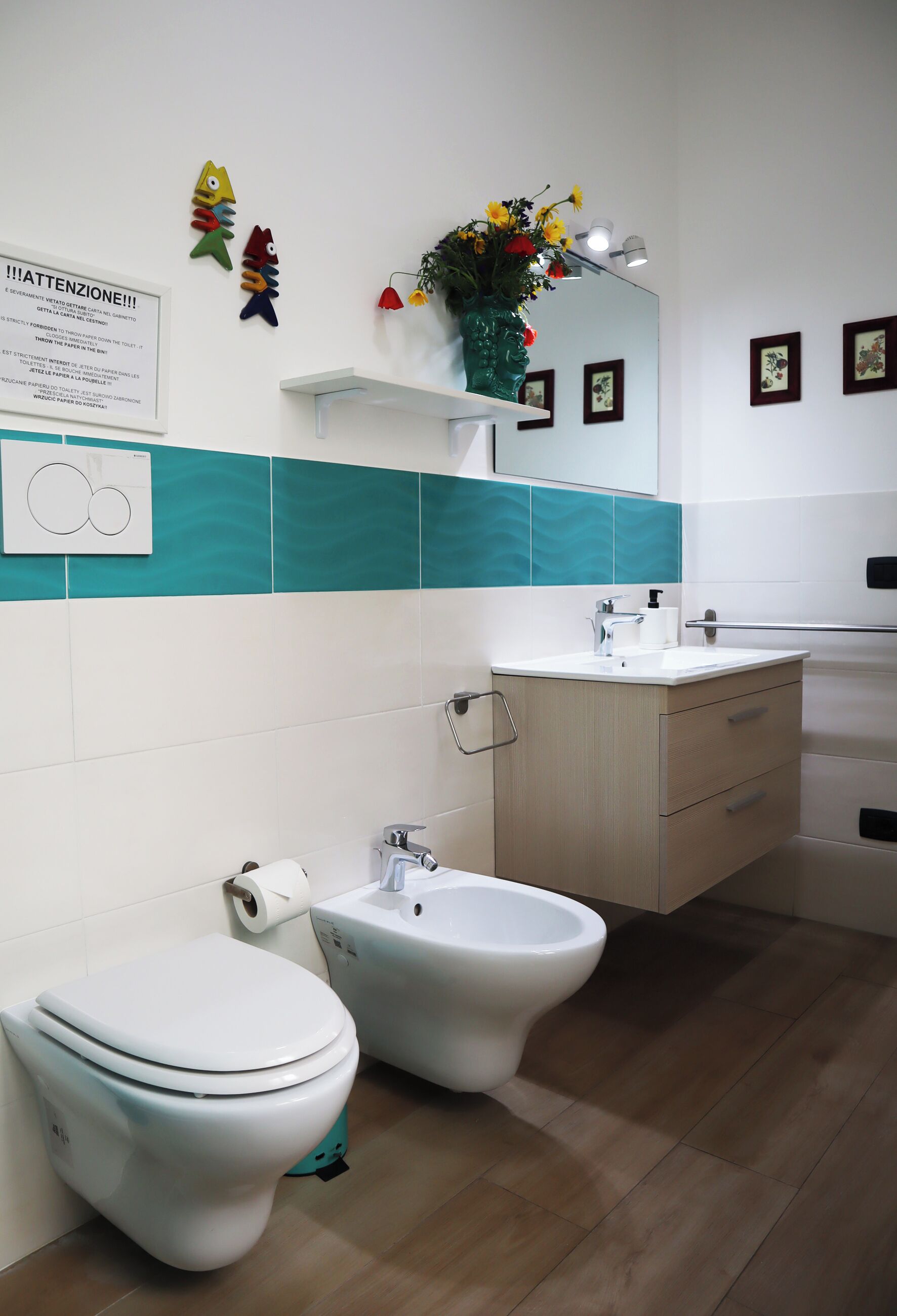 Et badeværelse med toilet, bidet, vask og spejl. Der er dekorative fisk på væggen, og et skilt med instruktioner.