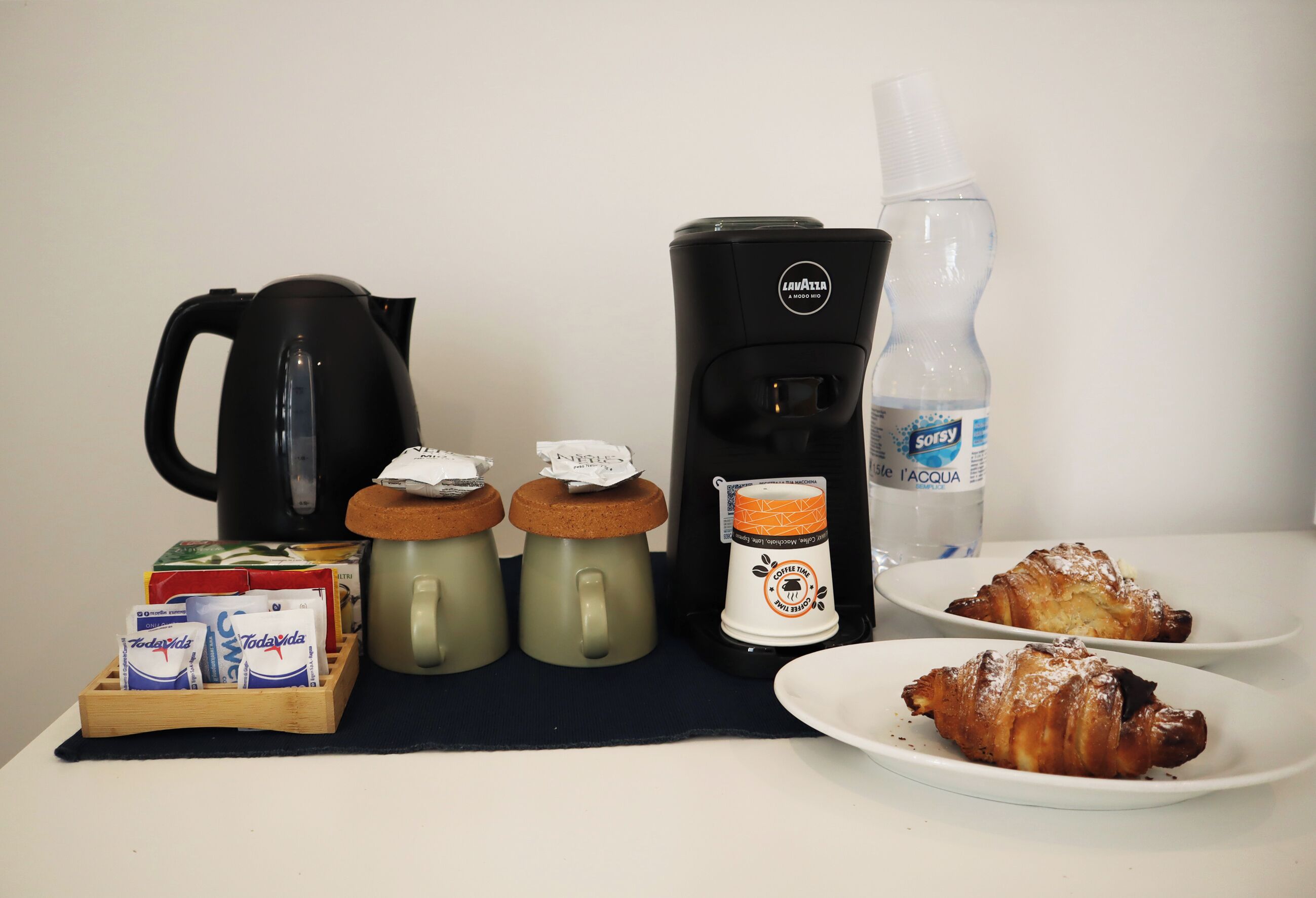 Preparación para el desayuno con cafetera, tetera, botella de agua y cruasanes sobre una mesa.