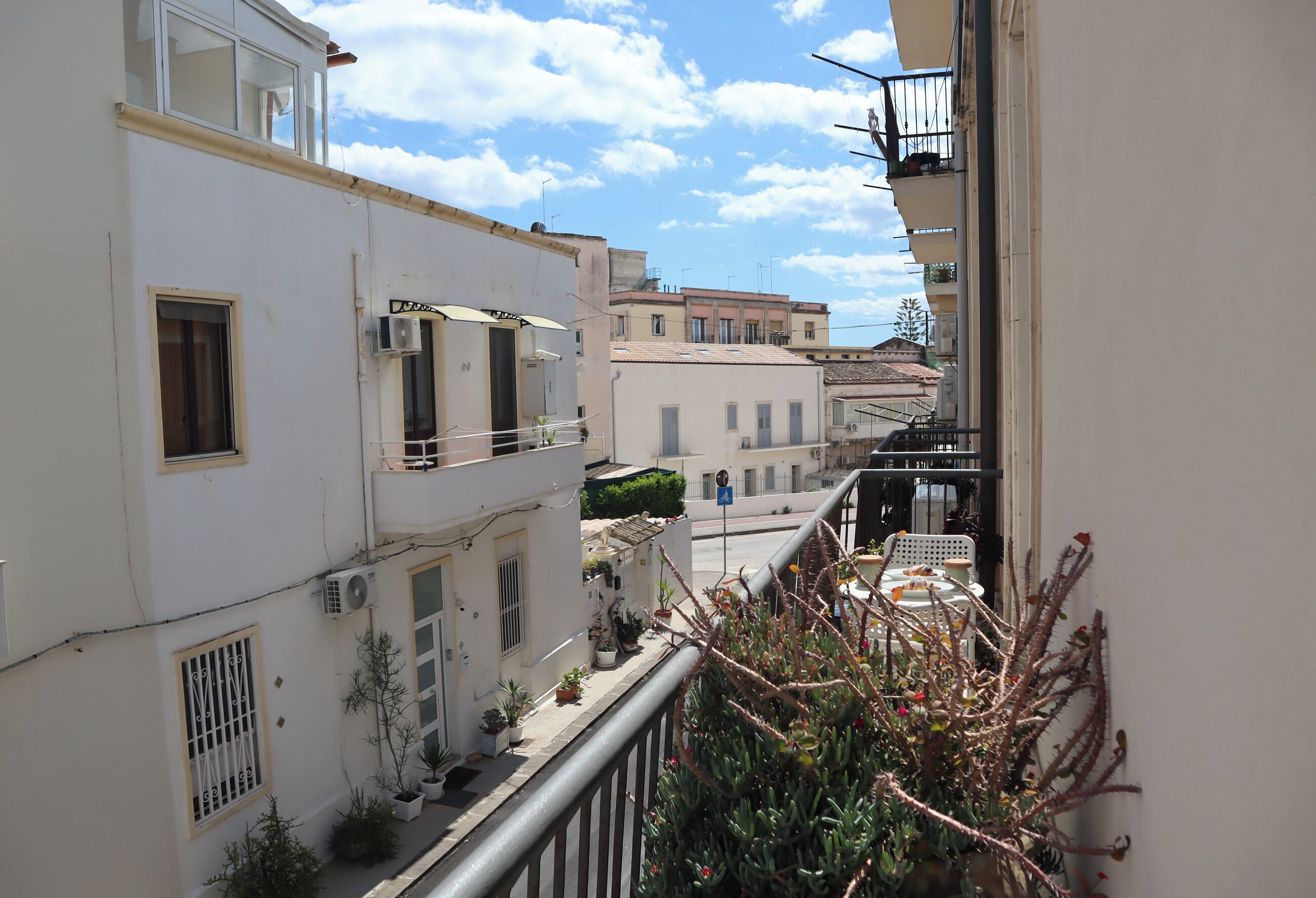 A narrow urban street with buildings, balconies, and potted plants.