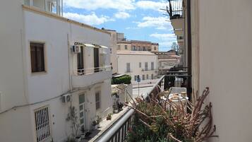 A narrow urban street with buildings, balconies, and potted plants.