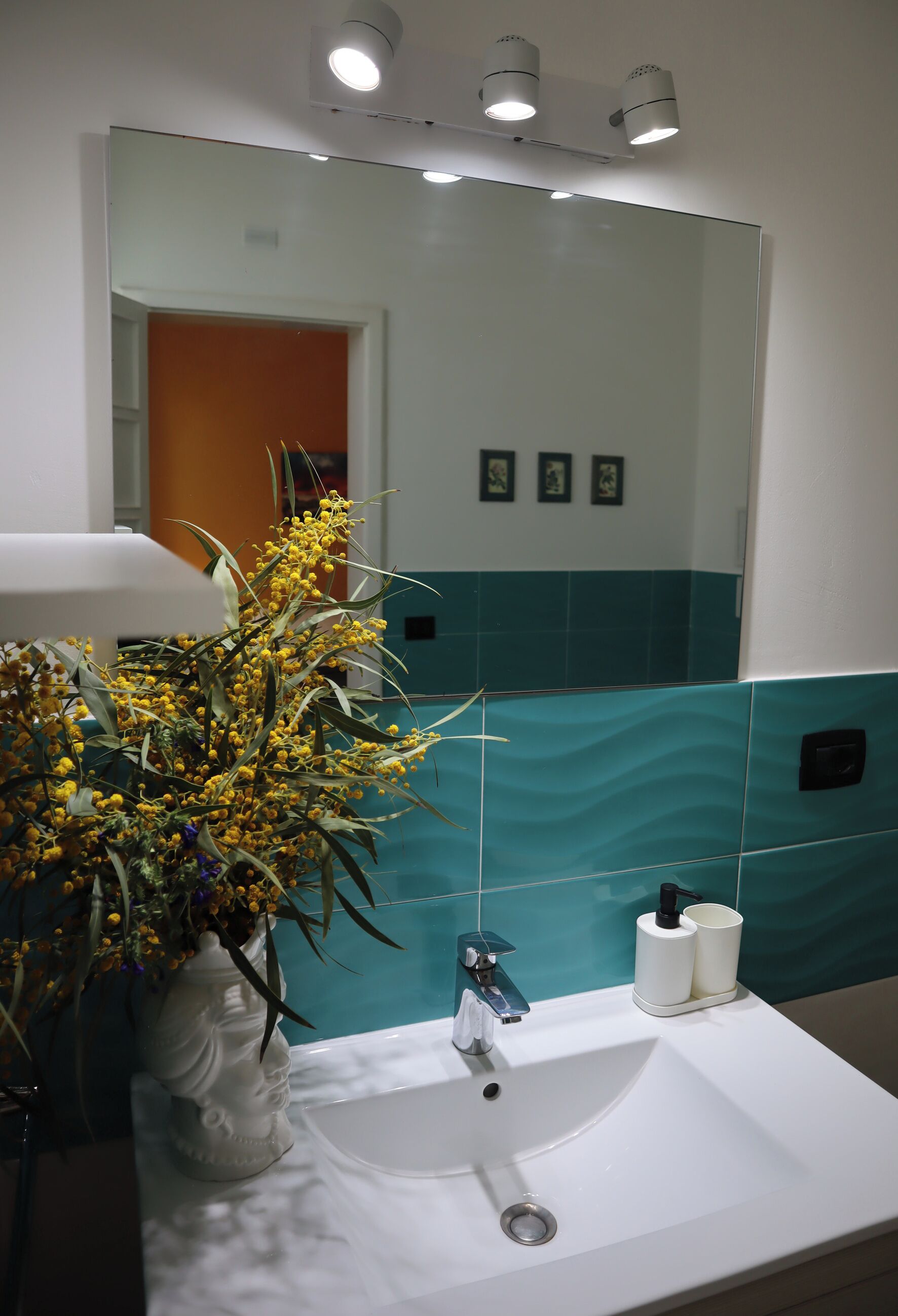 A bathroom with a white sink, a vase with yellow flowers, a mirror, and teal and white tiles.