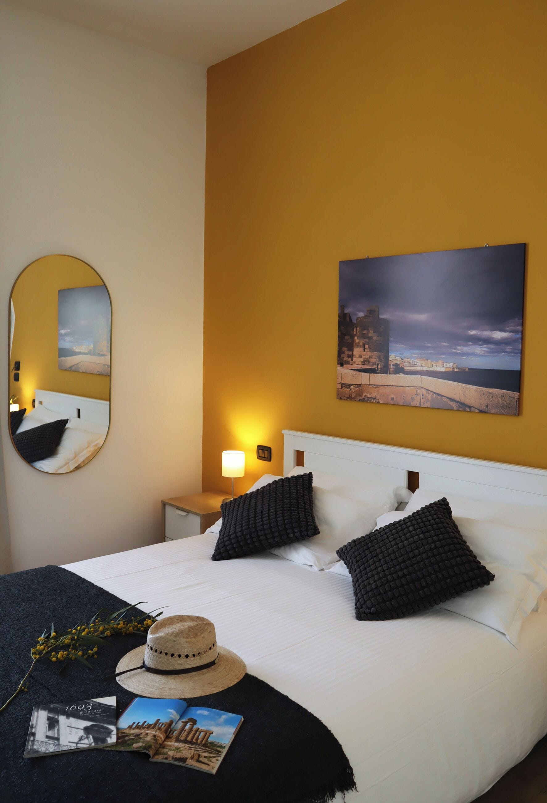 A neatly made bed with a white headboard, a mirror on the wall, a nightstand with a lamp, and a framed picture above the bed.