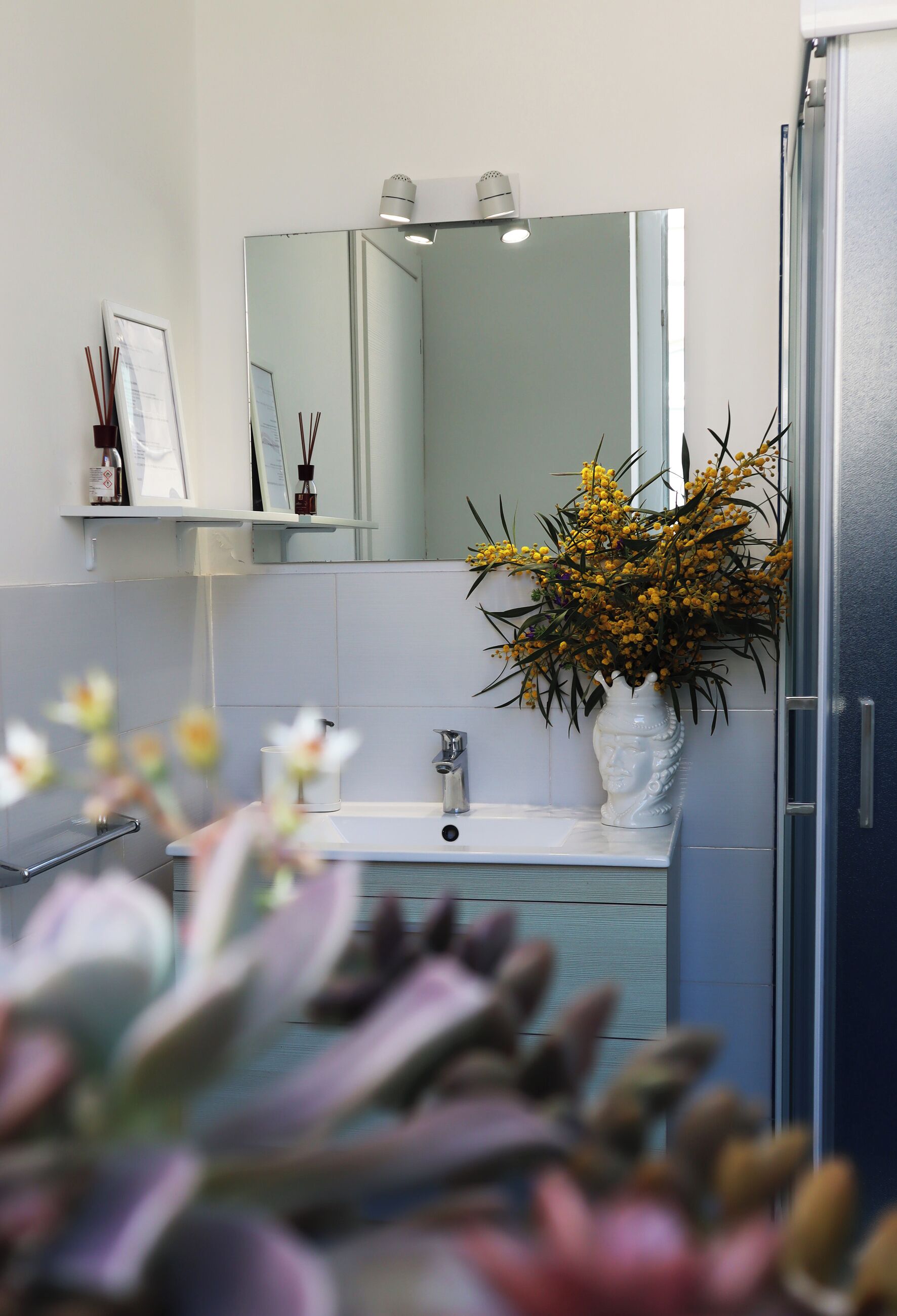 A bathroom with a white sink, a mirror above it, a vase with yellow flowers, and a shower door.