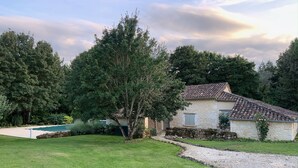 Une maison en pierre avec un toit de tuiles, entourée d’une pelouse bien entretenue et d’arbres.