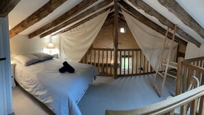 Une chambre avec un plafond haut, des poutres apparentes, un lit recouvert de draps blancs, une échelle et une vue à travers une fenêtre grillagée.