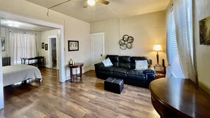 A spacious room with a ceiling fan, a black leather sofa, a wooden coffee table, a small side table, and a bed with a white headboard.