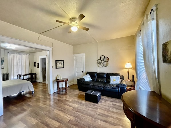 A spacious room with a ceiling fan, a black leather sofa, a wooden coffee table, a small side table, and a bed with a white headboard.