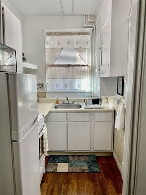 A compact kitchen with white cabinets, a stainless steel refrigerator, a sink, and a window with decorative curtains.