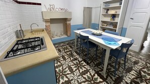 A compact kitchen with a gas stove, a sink, a 'Welcome' sign above the fireplace, a dining table with chairs, and a patterned floor. - Finished house, 100sqm 10 min from the beaches, 500 m from the historic center of Ugento (Ugento)