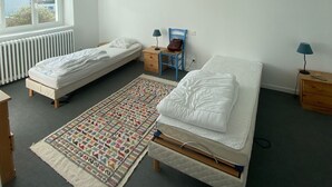 A bedroom with two single beds, a small table, a chair, a lamp, and a radiator.
