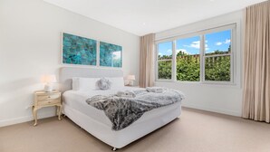 A bedroom with a large bed, two bedside tables, a window with curtains, and two abstract paintings on the wall.